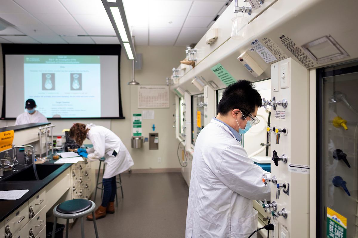 A smiling student works in an undergraduate chemistry lab while wearing a face mask and lab protective gear, including lab coat and goggles.